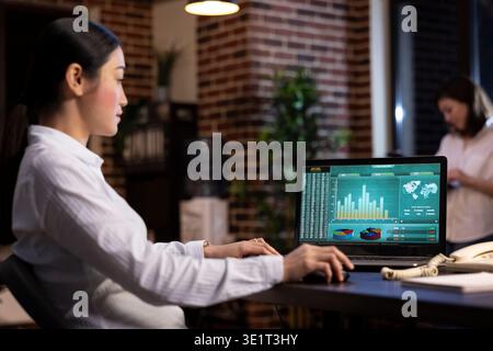 Businesswoman sitting in brick wall office, concentrated on analyzing marketing trends and company data on laptop as she works late at night. Professional analyst looking at charts displayed on screen. Stock Photo