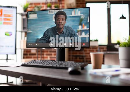 Closeup of computer monitor with virtual meeting in progress on empty desk in startup office. Black man seen on desktop pc in videoconference call as digital screen in background shows analysis data. Stock Photo