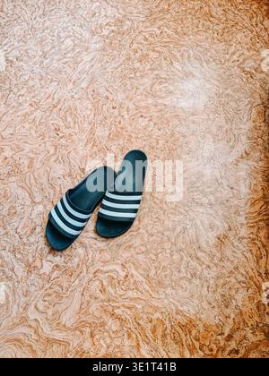 Top view of black slide sandals with white stripes placed on a textured marble floor. Clean minimal composition ideal for fashion, casual lifestyle, s Stock Photo
