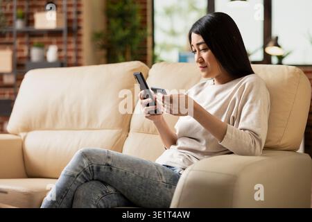 Focused businesswoman seated on couch, typing on cellphone, adding card information for online banking. Asian female manager remotely managing reservations and digital payments on mobile device. Stock Photo