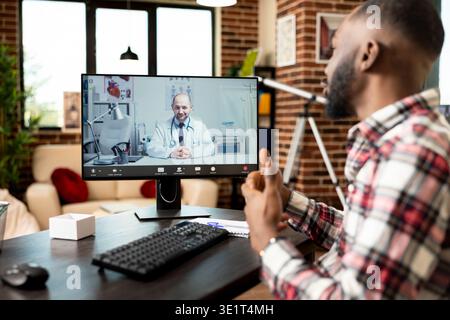 African american self employed man sits at home office desk, having virtual meeting with white male physician on desktop pc. Medical video call focuses on health advice and potential treatment options Stock Photo