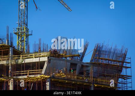 Construction workers install rebar and formwork on multi story concrete building, tower crane visible against clear blue sky. Stock Photo