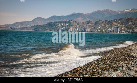 A professional wide-angle shot of a picturesque coastline. The image features a textured pebble beach in the foreground, white surf from breaking wave Stock Photo