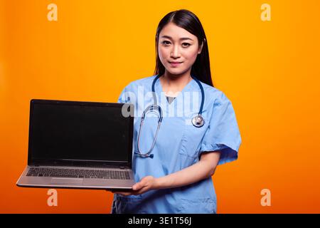 Asian nurse with stethoscope, carrying laptop with an empty display as she stands in front of isolated backdrop. Female healthcare worker in blue scrubs is holding a digital device with blank screen. Stock Photo