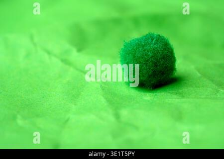 A small green pom-pom sits alone on crumpled green paper, creating a playful scene. Stock Photo