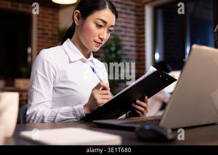Dedicated businesswoman with a pen in hand, reviewing documents on a clipboard in brick wall office. The modern desk setup, warm lighting, and laptop creates a productive work atmosphere. Stock Photo