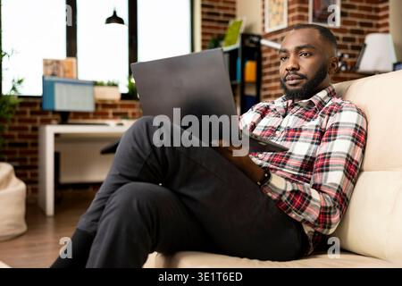 Remote worker sitting comfortably with legs crossed, using laptop to edit resume and search for new job roles. African american entrepreneur working on startup project files from home office. Stock Photo