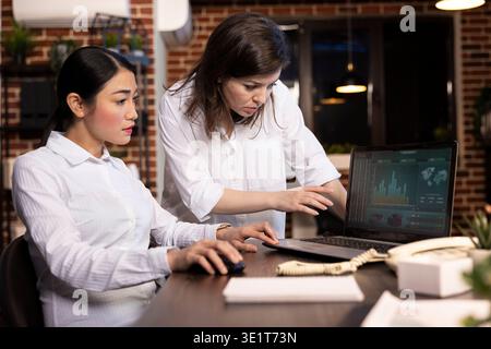 Diverse female colleagues work together at a desk, analyzing financial charts on laptop. Caucasian manager stands and explains details as her asian coworker listens attentively, engaged in discussion. Stock Photo
