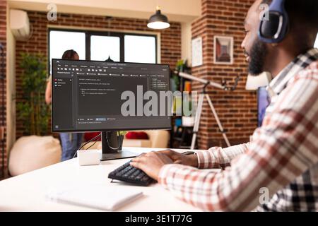 Remote male programmer sits at desk with headphones, coding and debugging scripts on computer. African american man listens to music and works on desktop pc, creating web application from home office. Stock Photo