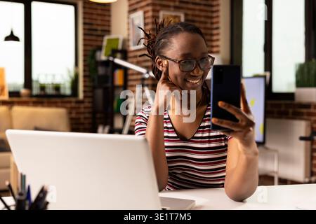 Smiling african american businesswoman seated at desk, having pleasant conversation on mobile device. Remote female employee taking break while using smartphone for video call in brick wall apartment. Stock Photo