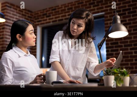Asian female executive with coffee in hand, collaborates on project with caucasian coworker, discussing business strategies. Businesswomen reviewing data on laptop during their night shift in office. Stock Photo