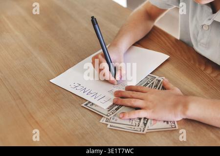 Close-up hands writing finance notes on paper with dollar bills on wooden desk, personal budget planning and money management concept, indoor lifestyl Stock Photo
