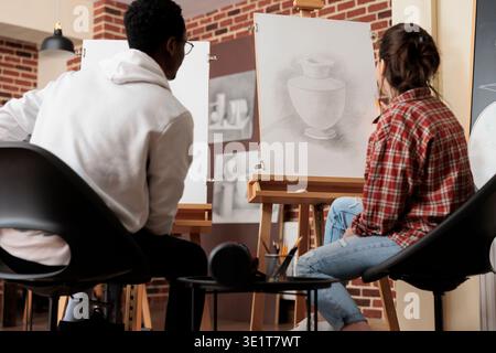 Young diverse people students looking at canvas with vase sketch, develop skills in line and shape while studying in art academy. Multiracial couple sitting at easels learning to draw together Stock Photo