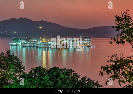 The Taj Lake Palace in Udaipur is brightly illuminated at dusk, creating vibrant light reflections on the calm surface of Lake Pichola. Stock Photo