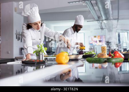 Female expert cook sprinkling tasty herbs to dish on the stove in kitchen, using fresh rosemary and spices to enhance the flavors and deliciousness while cooking the dish. Culinary arts. Stock Photo