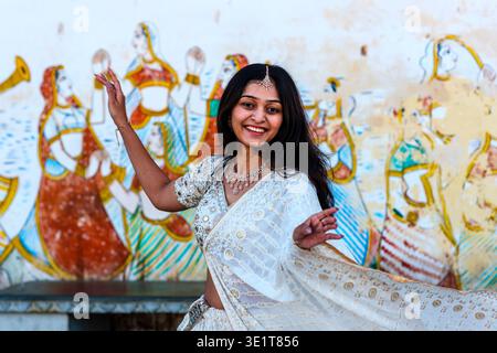A smiling woman in a white lehenga poses in Udaipur before a wall featuring traditional Rajasthani paintings of female figures and musicians. Stock Photo