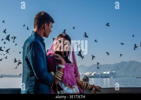 A couple in traditional attire looks at each other at Lake Pichola in Udaipur, with pigeons and the Taj Lake Palace visible in the distance. Stock Photo
