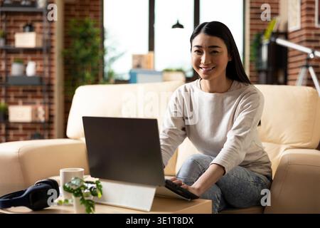 Cheerful female entrepreneur sitting confidently on sofa with laptop in home office. Smiling businesswoman working remotely, ready to review online articles to boost company progress. Stock Photo