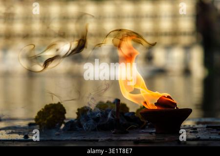 A lit diya with rising smoke and flames stands on a stone surface in Udaipur, with the blurred architecture of a lakeshore building in the distance. Stock Photo