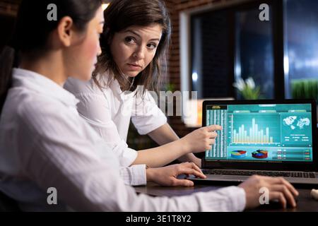 Two female colleagues collaborating on business analytics, reviewing data and discussing strategies on a laptop. They analyze financial information and plan tasks ahead in a professional workspace. Stock Photo