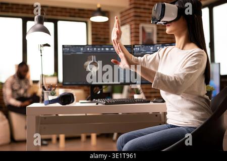 Female engineer wearing VR headset and gesturing hands in 3D simulation to review clamp model. Young woman works remotely with advanced technology, doing interactive engineering inspection on tools. Stock Photo
