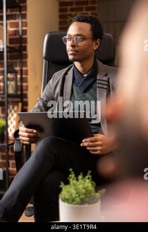 Psychiatrist sits in private clinic with laptop, listening and providing support to clients. Therapist wearing glasses conducts counseling session, helping patients cope with mental health challenges. Stock Photo