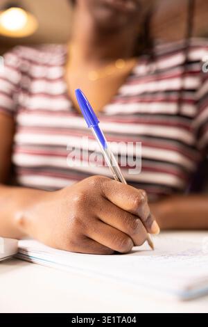 Selective focus on hand of female employee using pen to write down marketing strategy ideas on notepad. Close up of self employed black woman planning work schedule at home office desk. Stock Photo
