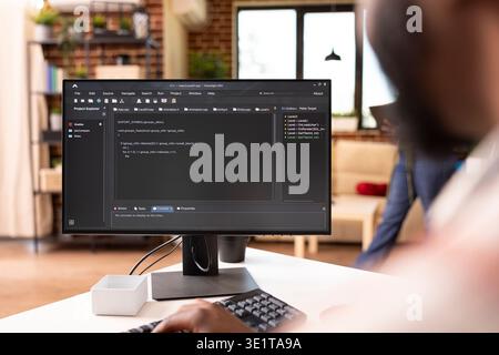 African american tech freelancer writes code on computer in modern home. Closeup of desktop monitor displaying programming scripts as black man builds and edits website from brick wall apartment. Stock Photo