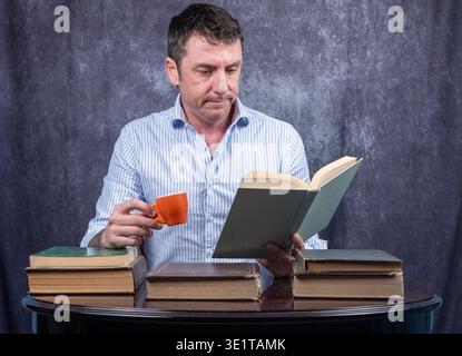 Man with a coffee cup in one hand and a book in the other. The man is sitting at a desk on which several books are placed and is wearing a shirt Stock Photo