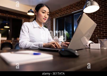 Corporate female professional engaged in her work, typing on a laptop at a modern office desk. Focused asian employee working on her personal computer, reviewing and updating business documents. Stock Photo