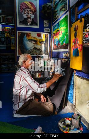 A local artist in Udaipur focuses on hand-painting a fabric piece, with several large portraits and traditional paintings displayed on the wall. Stock Photo