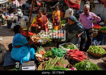 Customers interact with a seated vendor at a vibrant vegetable market in Udaipur, surrounded by carrots, radishes and green beans. Stock Photo