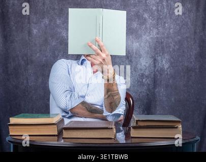 Man with face covered by the book he is reading. The man is wearing a white and blue shirt and is sitting at a desk with other books resting on top Stock Photo