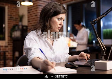 Closeup of caucasian woman working at her desk, focused on taking important notes on a notebook while using her laptop. The dim lighting highlights her night shift office environment. Stock Photo