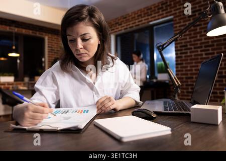 Focused female executive checking business reports on a clipboard at her desk. Dedicated businesswoman holding a pen and reviewing financial documents in a brick wall office. Stock Photo