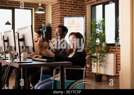 Smiling female employee, wearing wired headphones, works diligently at her desk. Caucasian woman in wheelchair uses her headset to communicate with online clients, entering data into her computer. Stock Photo