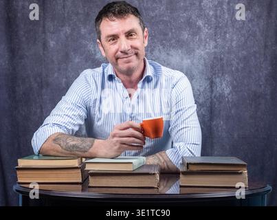 Man holding a coffee cup sitting at a desk on which several books rest. The man is forty-two years old and wears a white and blue striped shirt Stock Photo