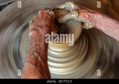 The mud-covered hands of a potter in Udaipur carefully shape a rotating piece of wet clay on a fast-moving wheel to create a traditional vessel. Stock Photo