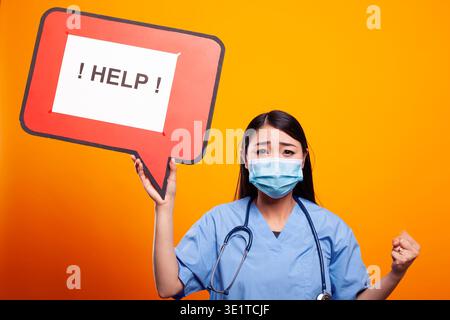 Asian woman doctor with her fist raised, asking for help using a cardboard speech bubble. Female nurse wearing scrubs and face mask, holding a red signboard against an isolated background. Stock Photo