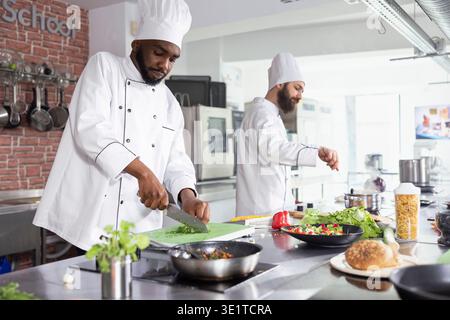 African american chef chopping herbs and spices on a cutting board, mincing raw ingredients for a fine dining dish. Young male cook using a big knife to slice fresh produce, gastronomy. Stock Photo