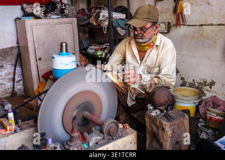 A craftsman in an Udaipur workshop uses a large industrial grinding wheel to sharpen a pair of metal scissors, creating a bright shower of sparks. Stock Photo