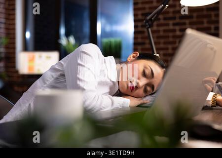 Overworked corporate manager sleeping at her office desk, taking a break during her night shift. Her laptop sits on the desk, highlighting the fatigue of long working hours in a modern workspace. Stock Photo