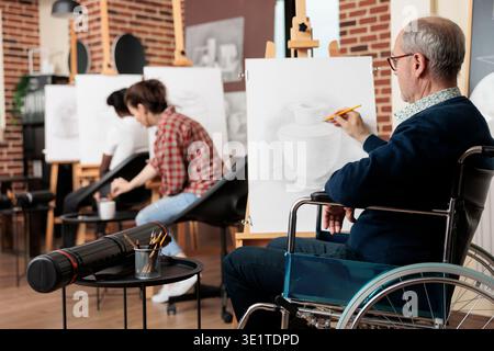 Senior man with physical disabilities sitting at easel during group art class, wheelchair user increasing fine motor skills through drawing practice. Disabled person engaging in creative activities Stock Photo