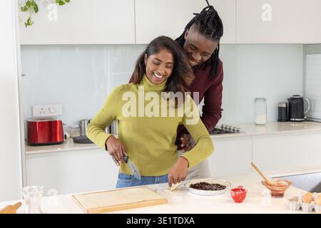 Diverse couple preparing pastry together at kitchen island, using bench scraper and pie dish Stock Photo