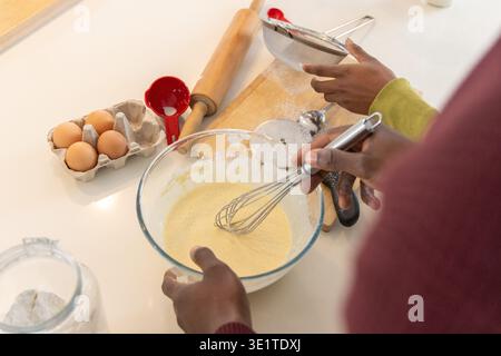 African American friends mixing batter in home kitchen using glass bowl, whisk, sieve, eggs Stock Photo
