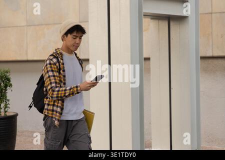Asian man walking on path beside building wearing beanie backpack holding phone and yellow folder Stock Photo