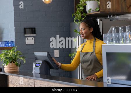 African American woman wearing mustard top and gray apron tapping POS at cafe counter, copy space Stock Photo