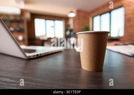 Closeup of disposable mug placed on vacant desk with laptop nearby, highlighting clean brick wall workspace. Coffee cup on table near digital device, set in startup office with no people. Stock Photo