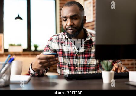 African american entrepreneur managing online transactions, reading credit card information at home office desk. Remote male manager updating financial data on computer in brick wall apartment. Stock Photo