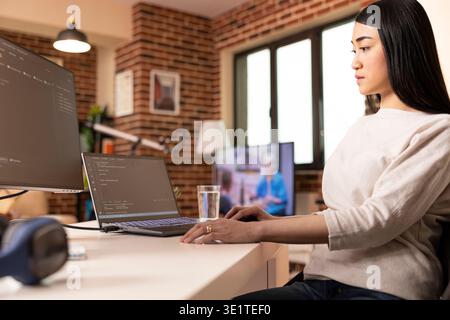 Asian software developer working remotely from home, programming across device screens. Female IT specialist seated at desk, reviewing coding scripts and maintaining online security. Stock Photo